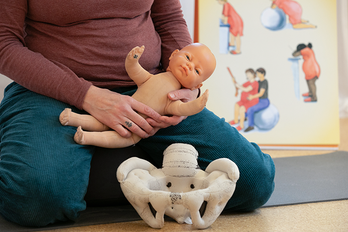 Eine Puppe wird in den Händen der Hebamme gehalten. Vor ihr liegt eine Nachbildung eines Beckens.